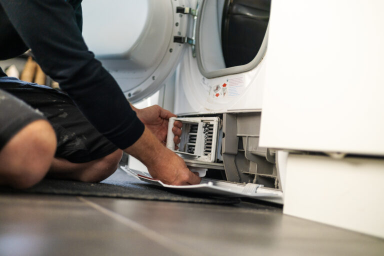 Person knæler foran åben tørretumbler og trækker snavset filter ud fra sockelåbningen.