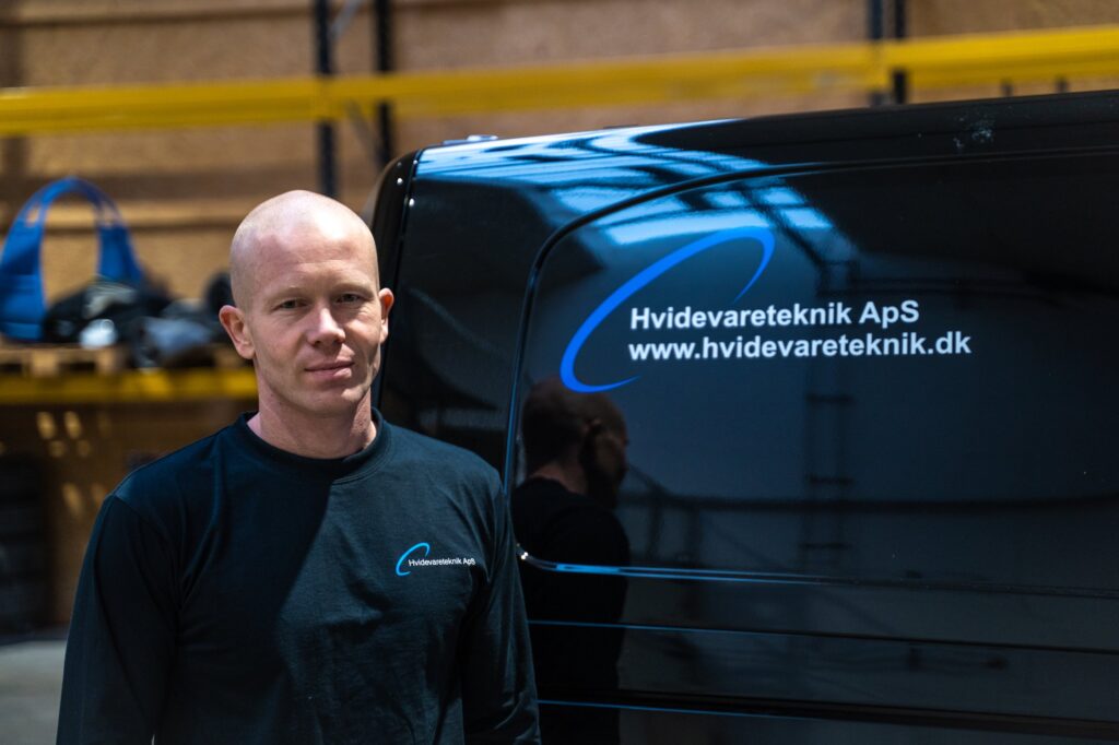 Montør fra Hvidevareteknik ved siden af en servicebil med firmaets logo.